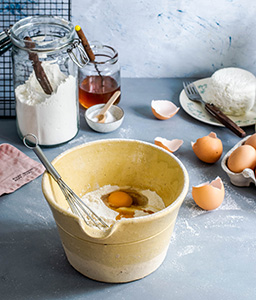 Scene of baking ingredients being mixed together. Brown egg shells, flour, and salt in background with mixing bowl with whisk, flour, and eggs inside. 